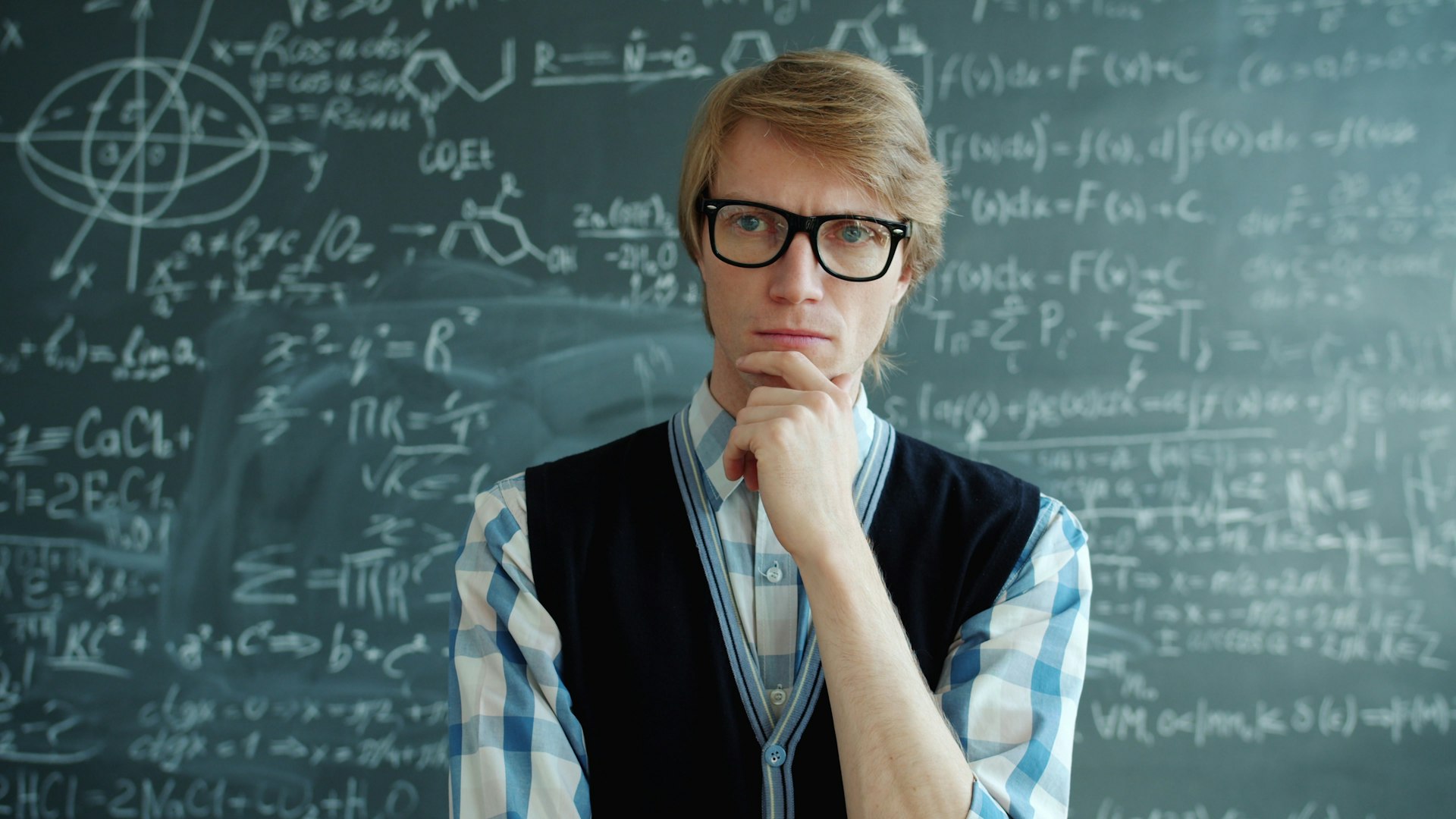 Man with glasses thinks in front of chalkboard.